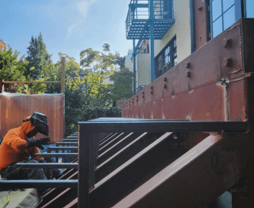 Construction worker in orange outfit welding metal beams next to a building under clear sky.