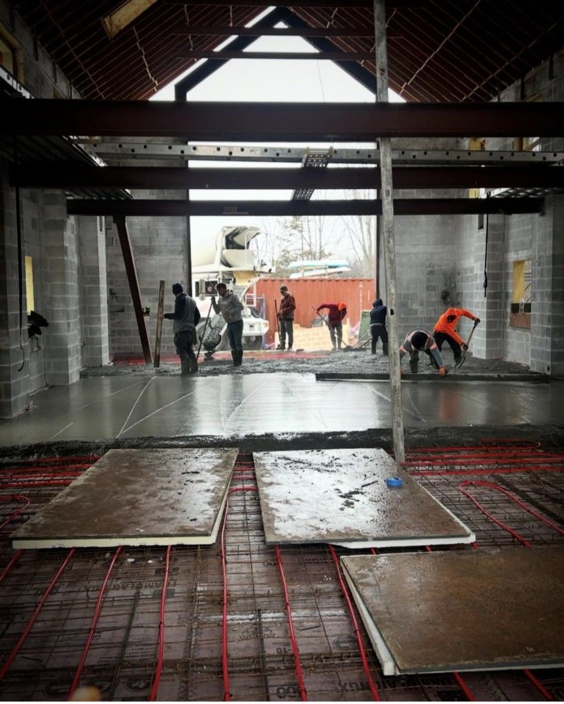 Construction workers pouring and leveling concrete inside a large building with exposed ceiling beams.