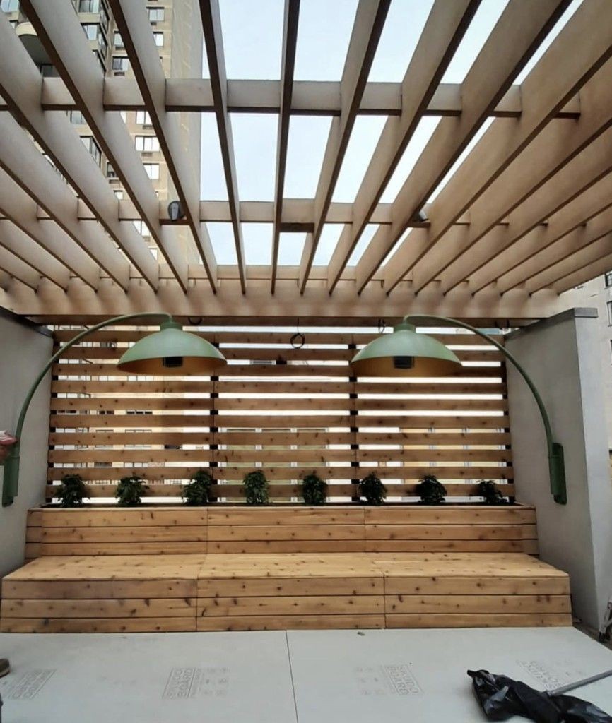 Outdoor seating area with wooden bench, pergola, and two large green lamps overhead.